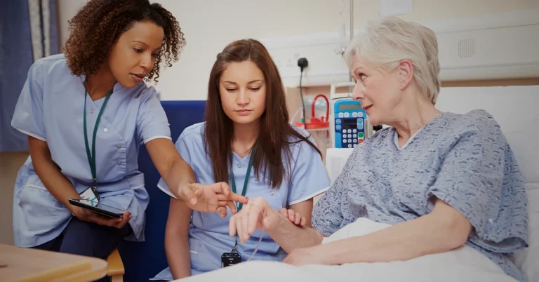 A trainee nurse learns from a nursing instructor during clinicals by a female patient’s hospital bed.