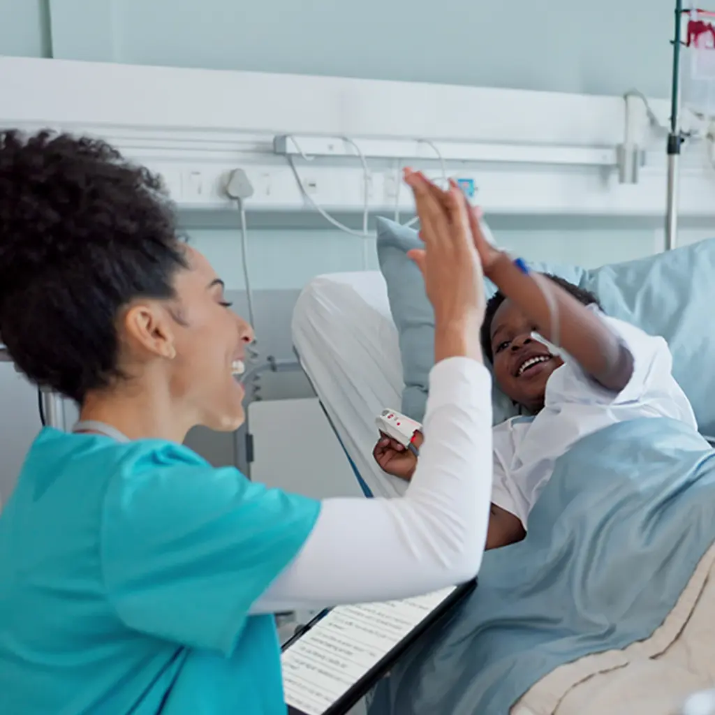 A female nurse shares a joyful high-five with a young child in a hospital bed.