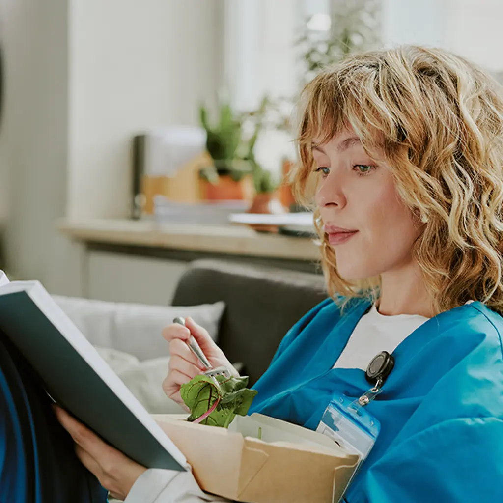 A nursing student in scrubs sits on a couch, studying and eating a salad.
