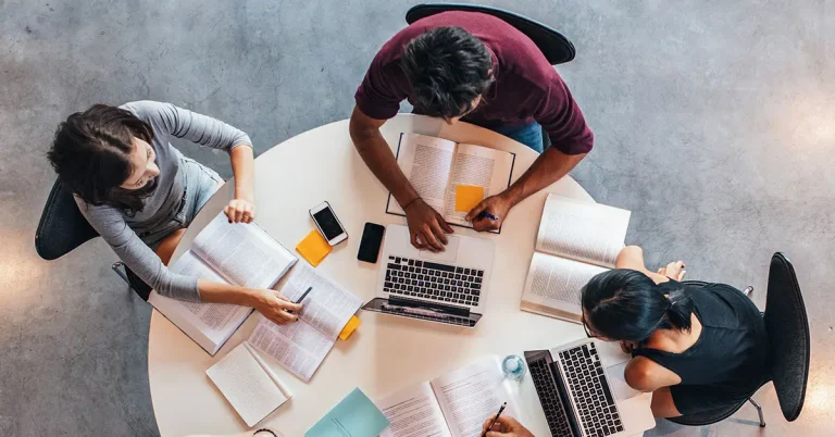 Top-down view of 3 students studying together with laptops, notebooks, and textbooks, collaborating on NCLEX prep.