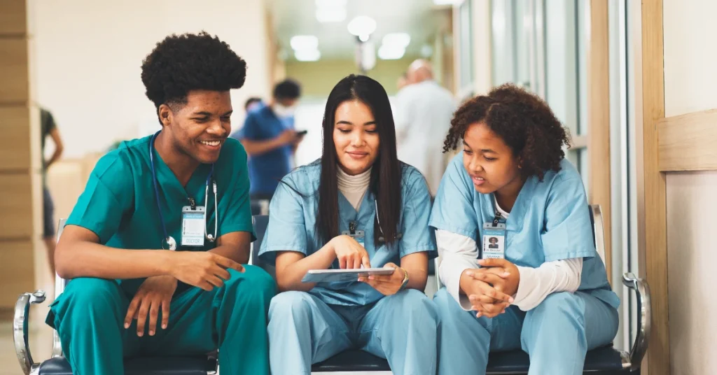 Three nurses huddle together in a hospital waiting room, reviewing notes.