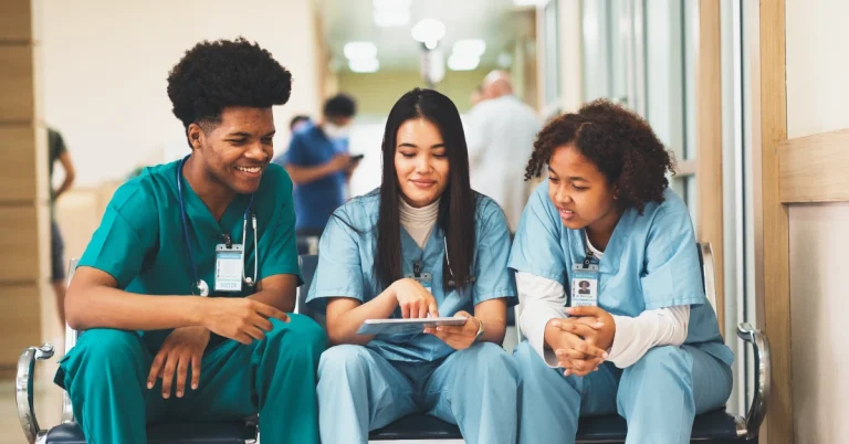 Three nurses huddle together in a hospital waiting room, reviewing notes.